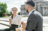 Man in black suit jacket sitting beside woman in white long-sleeve shirt holding brown paper cup