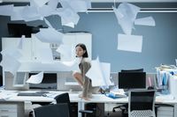 Woman crouching on a desk among flying papers in an office